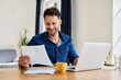 © baranq - Smiling man looking at financial document working remote on laptop