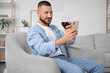 © Pixel-Shot - Young man with glass of wine using mobile phone in armchair on his day off at home