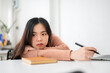 © bongkarn - Bored asian woman resting head on hand holding pen looking at laptop on table in classroom or office