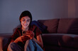 © we.bond.creations - Young woman relaxing at home, enjoying content on her mobile phone, with the screen illuminating her smiling face