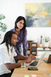 © Studio Marmellata - Diverse business professionals collaborate in a bright office. A man works on a laptop, and a woman stands beside him, smiling confidently at the camera.