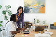 © Studio Marmellata - Diverse colleagues collaborate in a contemporary workspace. A man with dreadlocks works on a laptop, holding a pencil. A woman stands, leaning over, smiling at the camera