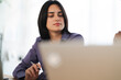 © Studio Marmellata - A young professional woman with dark hair works at her desk, focused on her laptop screen. She holds a pen, ready to write. This image shows concentration and productivity.
