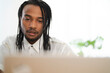 © Studio Marmellata - A young Black man with braided hair and a beard intently looks down at a laptop. He is engaged in work or study in a bright, modern setting.