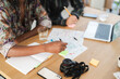 © Studio Marmellata - Two people collaborate at a wooden table. One writes in a notebook, the other uses a pencil. A map, laptop, camera, and smartphone are on the table, indicating planning or creative work.