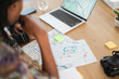 © Studio Marmellata - An individual plans a route or project at a desk, surrounded by paper maps, a laptop displaying a map, and sticky notes. A camera rests nearby.
