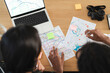 © Studio Marmellata - Two people collaborate on travel plans, examining physical maps and a laptop displaying a digital map. They are organizing their itinerary with sticky notes, preparing for an adventure.