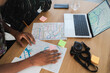 © Studio Marmellata - Two individuals are at a wooden table, studying maps and a laptop with a digital map. They are planning a journey, with sticky notes and a camera nearby for their adventure.