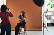 © Studio Marmellata - A female photographer captures a portrait of a male model posing on a stool in a professional studio. The model wears a patterned shirt and bandana. Studio lights are set up.