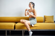 © Westend61 - Woman in athletic wear hydrating with water on yellow sofa at home