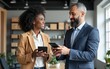 © Aria - Two happy busy diverse professional business team people talking standing in office using tab. Smiling young African woman employee and older Latin man executive having conversation at work.