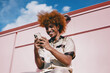 © BullRun - Smiling young Black man with curly red afro sitting outdoors, holding smartphone in both hands. Joyful moment of digital interaction under clear sky, expressing connection through technology.