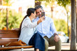 © Paula - Romantic couple taking a selfie while sitting close together on park bench