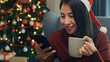 © tirachard - Positive Asia female wear red sweater new normal celebrate christmas smell aromatic coffee cup sit on wing chair check online greeting message from friend and family on phone in living room at house.