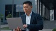 © Yuliia - Asian middle-aged businessman typing laptop in city Korean business man serious browsing wireless computer in downtown internet virtual job work task startup project Chinese Japanese male entrepreneur