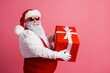 © deagreez - Santa in red suit with white beard wearing red glasses holds a large gift with white ribbon against pink background celebrating Christmas and holiday season