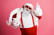 © deagreez - Santa in red suit with white fur and sunglass points up while smiling against a pink background celebrating christmas season