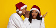 © Prostock-studio - A joyful couple is wearing red and white Santa hats. One person whispers in the other's ear, who looks surprised and excited. Their expressions capture the holiday spirit perfectly.