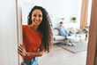 © Prostock-studio - A woman with curly hair stands at the door, smiling warmly at the camera. In the background, a man is seated comfortably on a couch, creating a relaxed home atmosphere.