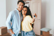© Prostock-studio - Two smiling individuals stand together in a bright living room filled with moving boxes. They proudly hold up a set of keys, celebrating their new home together.