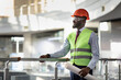 © Prostock-studio - Smiling african american young man in protective vest, helmet and glasses architect visiting building site, holding rolled drawing, looking at copy space, side view, panorama