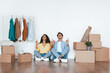 © Prostock-studio - A couple sits on the floor of their new home, surrounded by cardboard boxes. They smile and point upwards, expressing excitement about their fresh start and new beginning.