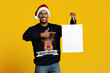 © Prostock-studio - A cheerful man in a Santa hat smiles broadly while holding an empty gift bag. He points to the bag, ready for Christmas celebrations. The background is a vibrant yellow, adding to the festive mood.