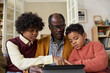 © Mediaphotos - Senior Black man sitting between two Black boys using digital tablet together, boys focusing on screen while man supervising, multigenerational family engaging with technology