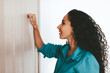 © Prostock-studio - A woman stands beside a wooden door, knocking with her fist. She has long, curly hair and wears a blue shirt. Her expression shows curiosity and anticipation as she waits for a response.
