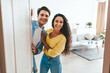 © Prostock-studio - A happy couple stands at the entrance of their stylish home. The man is wearing a blue shirt and the woman is in a yellow top. They are smiling warmly as they welcome visitors.