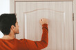 © Prostock-studio - A young man in a red sweater is knocking on a wooden door inside a well-lit room. The setting suggests a friendly visit or an invitation.