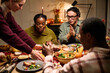 © DragonImages - Group of diverse young adults and Black woman sitting around table sharing Thanksgiving dinner, eating and talking, hands reaching for food, festive meal setting, autumn decorations visible