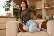 © Dexon Dee - Young woman holding her lower back with a painful expression, showing back pain while sitting at home.
