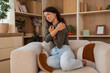 © Dexon Dee - Young woman holding her shoulder with a painful expression, showing shoulder pain while sitting at home.
