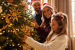 © Home-stock - Parents and daughter having fun decorating Christmas tree, placing Christmas lights and ornaments on it, decorating home for winter holiday season