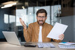 © Liubomir - Young man in glasses sitting at office desk feeling frustrated about business problems, holding up a document and throwing his other hand in the air in annoyance