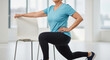 © SYATRI RAWU - Active senior woman in sportswear doing a forward lunge exercise using a chair for balance and support during her workout