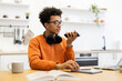 © sofiko14 - A young man with glasses speaks into his phone while working at a table in a modern kitchen setting.