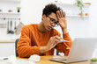 © sofiko14 - A young Black man with glasses checks his temperature while working on his laptop at home, appearing unwell.