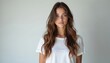 © miss irine - Young woman with long wavy brown hair stands against plain white studio background. Wears simple white t-shirt, looks directly forward with neutral expression. Soft indoor lighting illuminates face.
