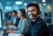 © miss irine - Young Middle Eastern man with headset works at computer in office. Customer service representative smiles at camera. Colleagues work behind in call center environment. Office workers provide support