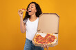 © Prostock-studio - Excited Latin young lady bites into a tasty slice of pizza while holding a carton box in a vibrant yellow-orange studio background. She showcases her love for junk food and pizza.
