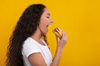 © Prostock-studio - Smiling female model bites into a tasty burger with excitement. She is in a casual outfit against a vibrant yellow wall. Her joyful expression shows her love for this delicious fast food snack.