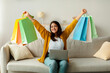 © Prostock-studio - An excited Arab woman sits on her sofa with a laptop, raising colorful gift bags above her head in happiness. She enjoys shopping online and is pleased with her purchases and discounts.