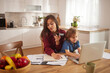 © Stockphotodirectors - A mother and her child are engaged in activities at a kitchen table. The mother speaks on a phone while working on a notebook, and the child types on a laptop nearby.