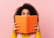 © SAYERA - Young woman with curly hair peeking over orange book, surprised