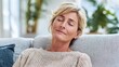 © Oleksandr - Middle-aged woman is relaxing on the couch with her eyes closed. Serene expression on face as she rests. A modern, calming scene indoors with soft, natural lighting.