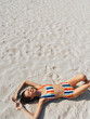© SHOTPRIME STUDIO - Young woman lying on white sand beach in a colorful striped bikini, sunlit and relaxed, capturing a carefree summer moment with warm tones and gentle breeze.