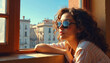 © Viktor - Young woman wearing sunglasses leans on windowsill looking at city buildings. Sunlight illuminates her face as she gazes outside, lost in thought or daydreaming.