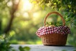 © Olya - Wicker picnic basket with red and white checkered cloth and fresh fruit on a wooden table beneath sunlit green trees, peaceful summer morning